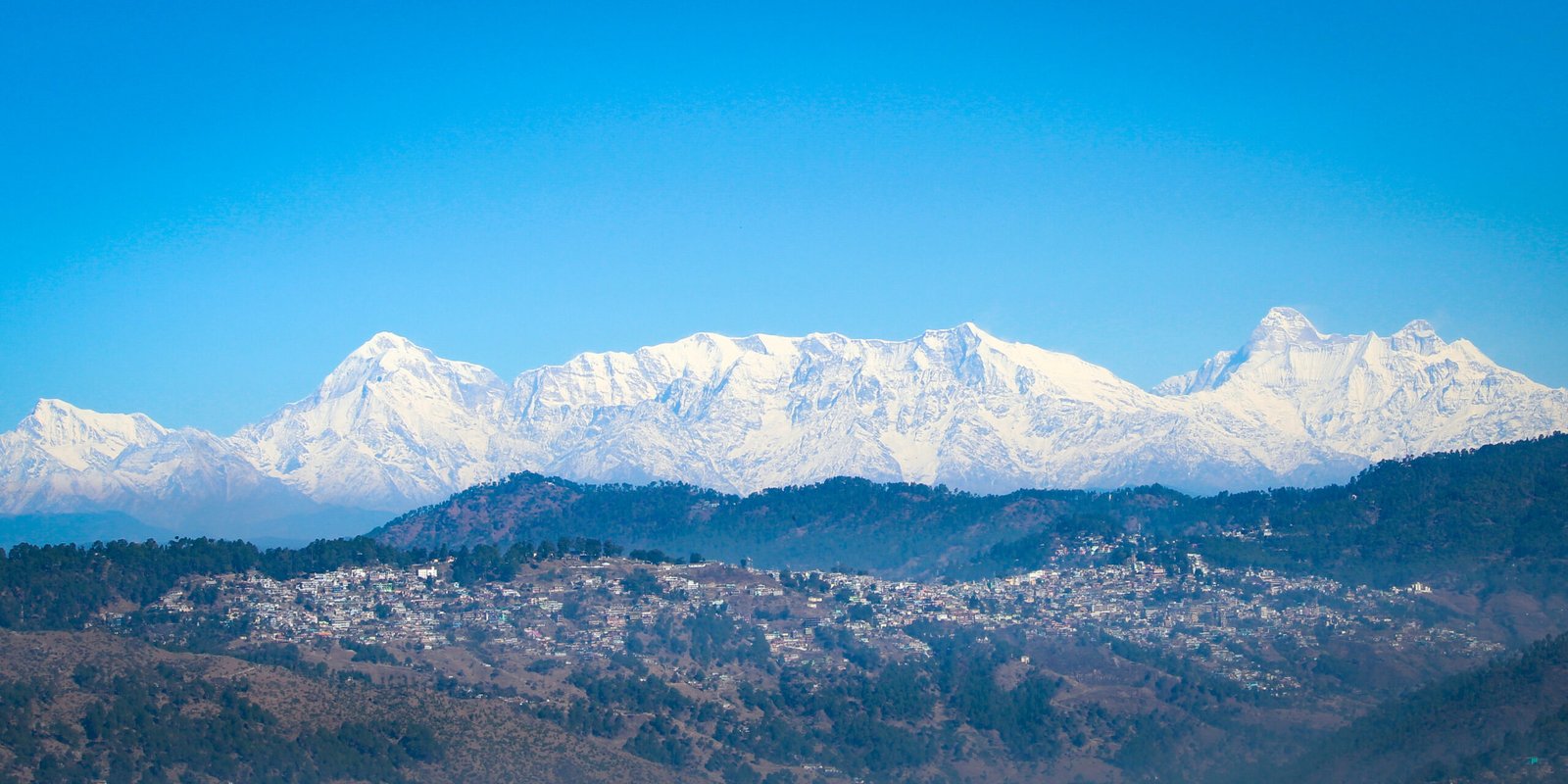 Layers of forested hills with the town of Almora spread across a ridge in the middle distance, and a row of snow-covered Himalayan peaks along the horizon under a clear blue sky.