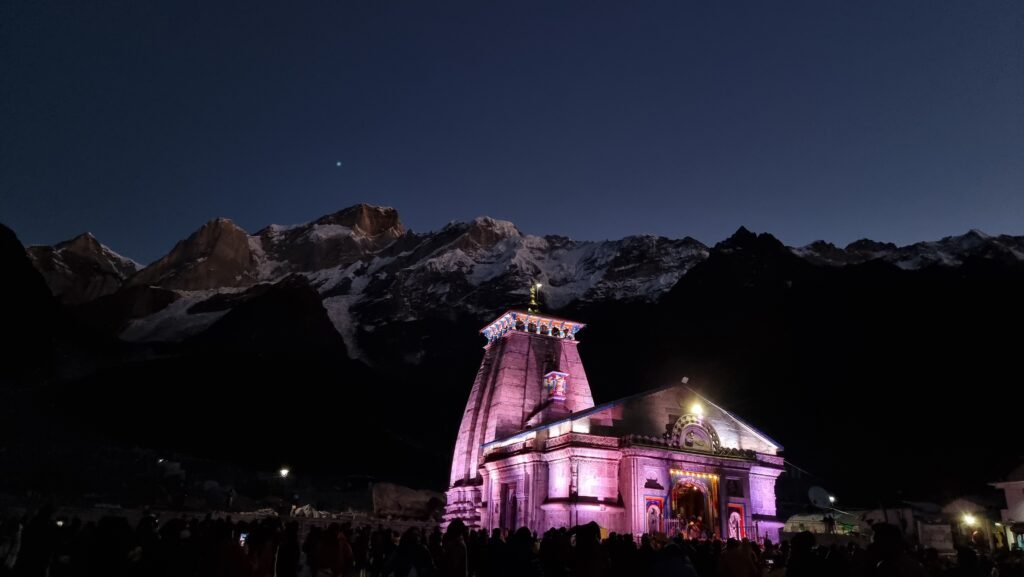 The Kedarnath temple at night, illuminated in pink and purple light, with dark Himalayan peaks rising behind it and a dense crowd of pilgrims gathered at the base of the temple.