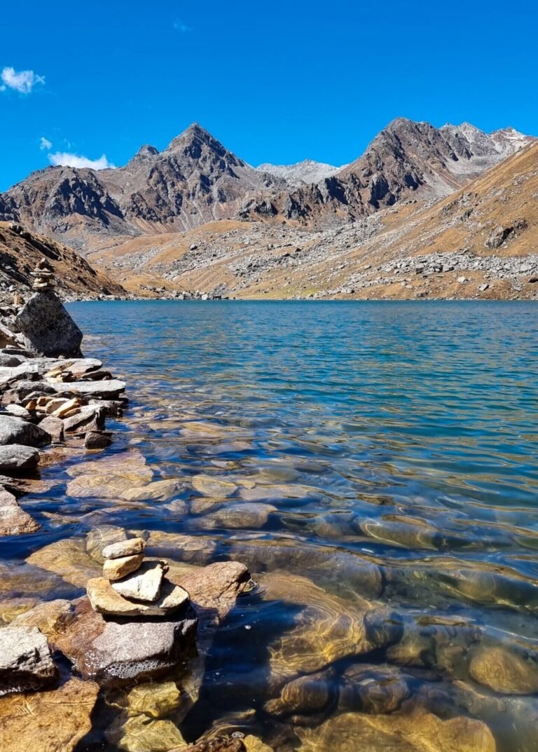 A clear blue high-altitude lake with stones visible under the water near the shore, small stacked stone cairns on the rocky bank, and bare brown mountain ridges under a deep blue sky.