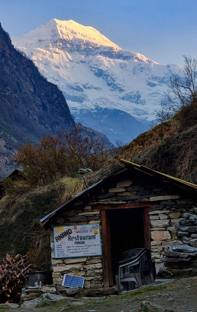 A stone hut with a hand-painted restaurant sign listing meals, with plastic chairs and a small solar panel outside. Behind it, a snow-covered Changuch peak lit by early morning sun above a dark valley.