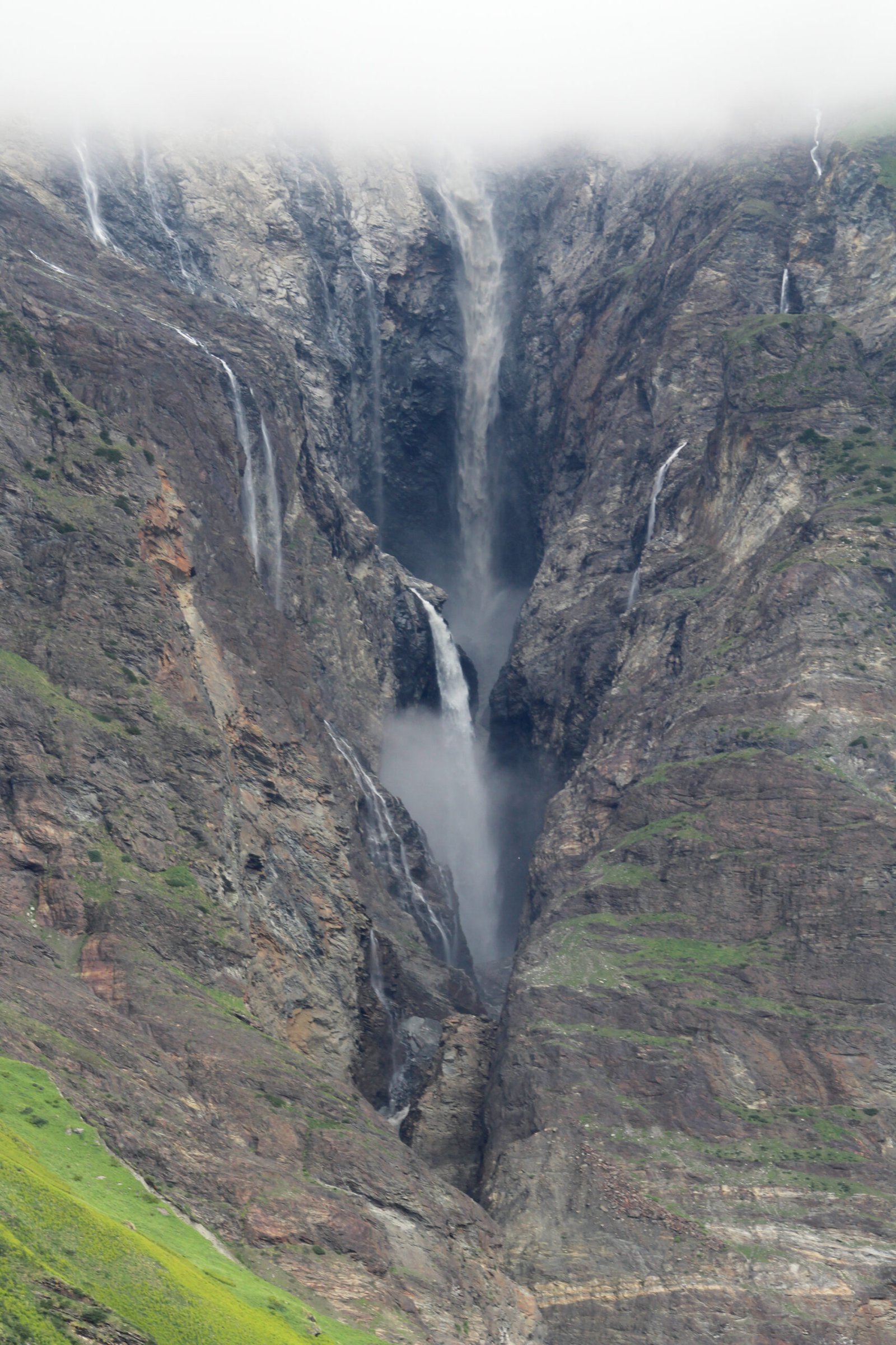 Multiple waterfalls streaming down a steep gray rock face into a narrow gorge, the tallest fall emerging from low cloud at the top of the frame, with green slopes at the lower left.