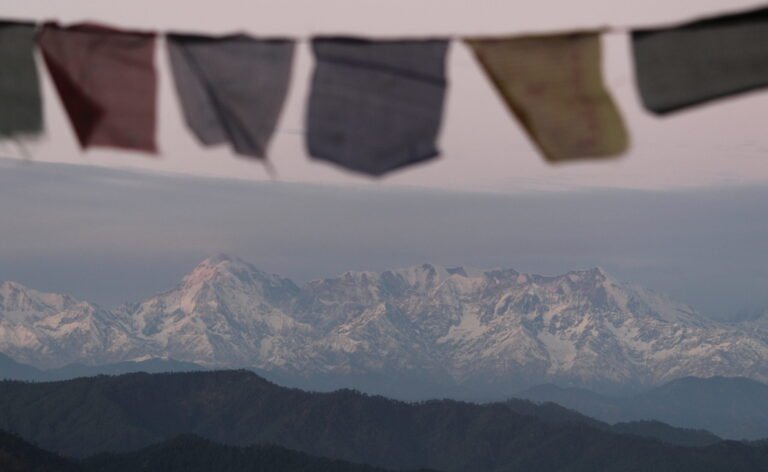 Five prayer flags hanging on a line in the foreground, out of focus, with snow-covered Himalayan peaks in sharp focus behind them under a pale evening sky, dark forested ridges below.